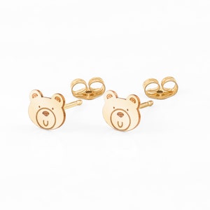 May include: A pair of gold bear stud earrings. The bear is outlined in a darker gold and has a smiling face.