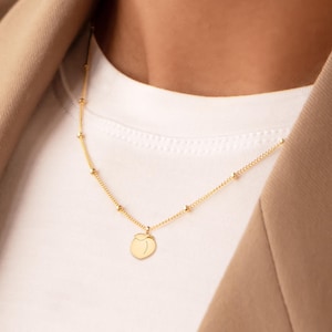 May include: A gold necklace with a peach pendant. The necklace has a fine chain with small, evenly spaced beads. The pendant is a simple, round peach shape. The necklace is worn over a white t-shirt and a beige blazer.