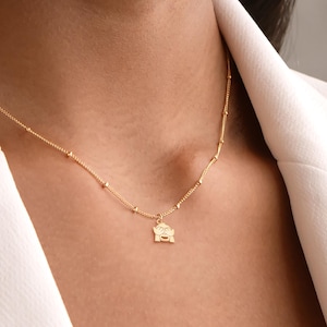 May include: A gold chain necklace with a small gold pendant featuring a stylized face with closed eyes. The necklace is worn by a person with a white blazer.