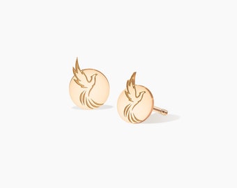 May include: A pair of gold-colored stud earrings. Each earring features a circular design with an engraved image of a dove in flight. The earrings are displayed on a white background.
