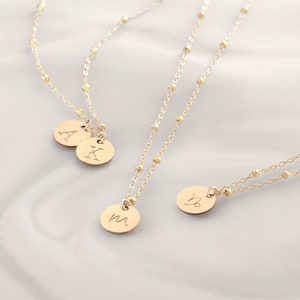 May include: Three gold necklaces with initial charms. The necklaces feature a delicate chain with small gold beads. The charms are round and feature the letters A, K, M, and the astrological symbol for Virgo.