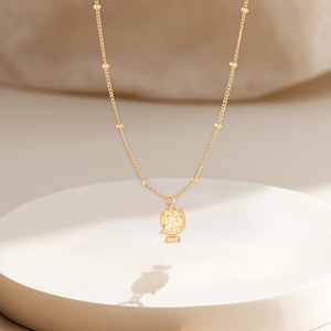 Może przedstawiać: Złoty naszyjnik z zawieszką w kształcie globu. Naszyjnik ma delikatny łańcuszek z małymi, równomiernie rozmieszczonymi koralikami. Zawieszka w kształcie globu to szczegółowy kontur Ziemi, zamontowany na podstawie. Naszyjnik jest prezentowany na białej, okrągłej platformie.