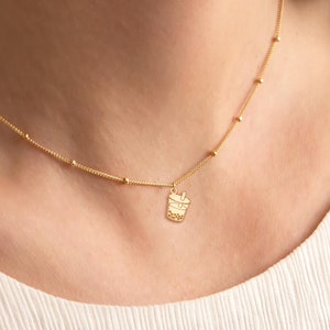 Puede incluir: Un collar de oro con un pequeño dije de oro en forma de taza de boba. El collar tiene una cadena delicada con pequeñas cuentas redondas.
