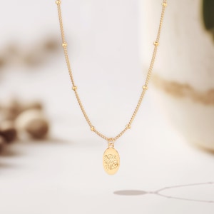 May include: A delicate gold-colored necklace with a small oval pendant. The pendant has an engraved design and the chain features small, evenly spaced beads. The necklace is displayed against a soft, blurred background.