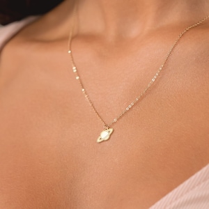 Peut inclure: Un délicat collier doré avec un petit pendentif en forme de planète. Le pendentif a un centre blanc et un anneau autour. Le collier est porté autour du cou, un bijou simple et élégant.
