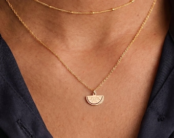 May include: A gold-toned layered necklace set consisting of two chains. The shorter chain features small evenly spaced beads, while the longer chain includes a small, flat, semi-circular pendant engraved with a sunburst pattern.
