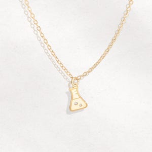 May include: A gold-colored necklace featuring a pendant shaped like a laboratory flask. The flask pendant has a white enamel finish with three small, clear accents. The chain is delicate and has a link design.