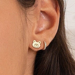 May include: A pair of gold earrings, one featuring a small bear design and the other a small hoop with sparkling stones.
