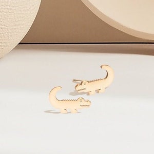 Könnte beinhalten: Ein Paar goldfarbene Alligator-Ohrringe. Die Ohrringe sind klein und zart und haben ein realistisches Alligator-Design.