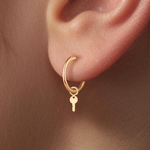 May include: Gold hoop earrings with a small key charm dangling from the bottom. The earrings are worn in the earlobe.