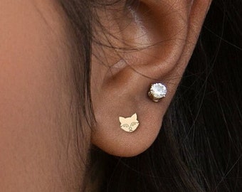 May include: A close-up of a person's ear with two earrings. The left earring is a small gold cat face, and the right earring is a small silver round stone.