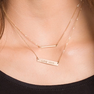 May include: A gold necklace with two chains. The bottom chain has a bar pendant with the words "Love Mom" engraved on it. The top chain has a plain bar pendant.