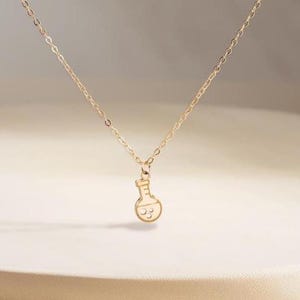 May include: A delicate gold-colored necklace featuring a pendant shaped like a laboratory flask with a smiling face. The necklace hangs on a thin chain, displayed against a neutral background. The pendant is approximately 0.5 inches in height.