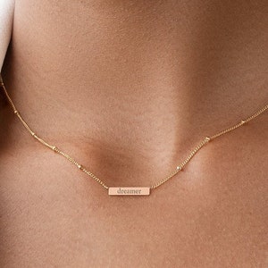 Peut inclure: Un collier en or avec une chaîne délicate et un petit pendentif en forme de barre qui dit "dreamer".