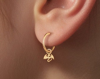 May include: Gold hoop earrings with a small dangling charm shaped like a bat. The earrings are worn in the earlobe of a person with dark hair.