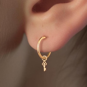 May include: Gold hoop earrings with a small key charm dangling from the bottom. The earrings are worn in the lower lobe of the ear.