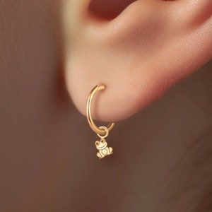Pode incluir: Um close-up de uma orelha com dois brincos de ouro. O brinco no lóbulo superior é um pequeno brinco de coração. O brinco no lóbulo inferior é um aro com um pequeno charme de sapo de ouro pendurado.