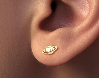 Puede incluir: Primer plano de una oreja con un pequeño pendiente de oro en forma de planeta Saturno. El pendiente tiene un centro blanco y un fino anillo de oro alrededor.