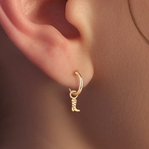 May include: Gold hoop earrings with a small gold boot charm hanging from one earring. The earrings are worn in a person's ear.