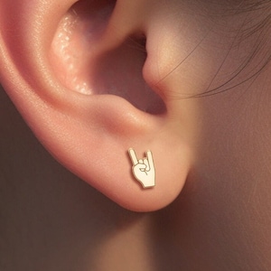 Könnte beinhalten: Nahaufnahme eines goldfarbenen Ohrsteckers in Form einer Hand, die das Rock'n'Roll-Zeichen macht. Der Ohrring befindet sich an einem Ohr.