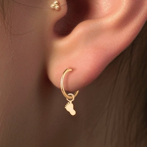 May include: A person's ear with three gold earrings. The top earring is a small cluster of gold beads. The middle earring is a gold hoop. The bottom earring is a gold hoop with a small gold footprint charm hanging from it.