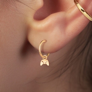 May include: Gold hoop earrings with a small gold charm in the shape of a video game controller hanging from one earring. The earrings are worn in a person's ear.
