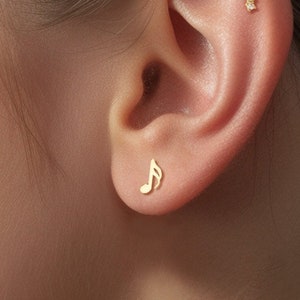 Könnte beinhalten: Nahaufnahme eines Ohres mit zwei goldenen Ohrringen. Der linke Ohrring ist eine kleine Musiknote, der rechte Ohrring ist eine gebogene Stange mit fünf kleinen Diamanten.