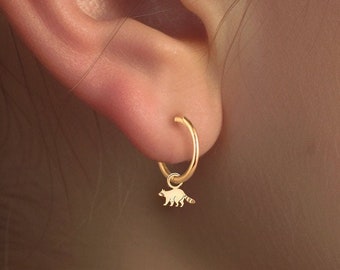 Könnte beinhalten: Goldfarbener Ohrschmuck, bestehend aus einem Mond-Ohrstecker mit kleinen Edelsteinen, einem schlangenförmigen Helix-Piercing und einem Creolen-Ohrring mit einem Waschbär-Anhänger. Der Schmuck wird an einem Ohr präsentiert.