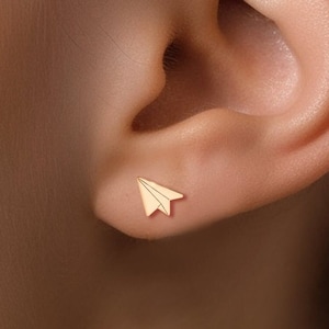 以下が含まれることがあります： 金色の紙飛行機のイヤリングをした耳のクローズアップ。