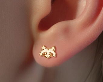 Puede incluir: Un par de pendientes de color dorado con un diseño de cara de mapache. Los pendientes son pequeños y delicados, perfectos para el uso diario.