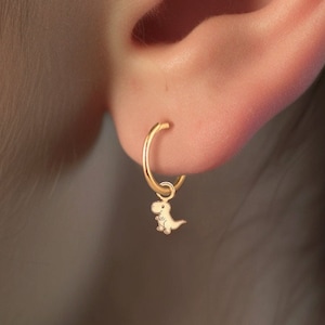May include: Gold hoop earrings with a small dinosaur charm hanging from one earring. The earrings are worn in the earlobe and the helix piercing.