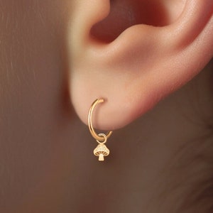 Puede incluir: Aretes de aro de oro con un pequeño dije de hongo que cuelga de uno de los aretes. Los aretes se usan en el lóbulo de la oreja.