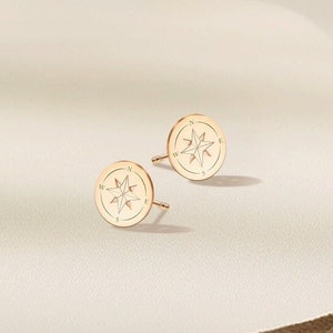 Puede incluir: Un par de pendientes de oro en forma de brújula con un diseño de estrella. Los pendientes son pequeños y delicados.