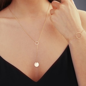 Puede incluir: Un conjunto de joyas doradas que incluye un collar y una pulsera. El collar presenta un colgante circular y una cadena larga con un pequeño círculo y un colgante de disco. La pulsera tiene una cadena delicada con un dije circular.