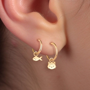 May include: A pair of gold hoop earrings with small charms. The left earring has a fish charm and the right earring has a cat charm.