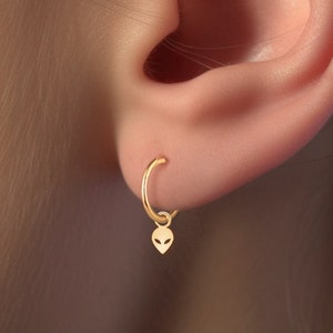 May include: A gold hoop earring with a small alien charm dangling from it. The earring is worn in the left earlobe.