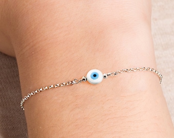 Puede incluir: Un brazalete de cadena de plata con un pequeño amuleto de ojo turco azul y blanco.