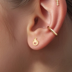 Puede incluir: Primer plano de la oreja de una persona con tres pendientes de oro. El pendiente inferior es un pequeño diseño de llama, el pendiente del medio es un aro y el pendiente superior es una fila de pequeños diamantes.