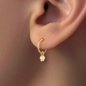 Könnte beinhalten: Goldene Creolen mit einem kleinen Hamsa-Hand-Anhänger, der an jedem Creolen hängt.