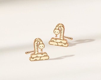 May include: A pair of gold-colored stud earrings. Each earring features a stylized snake design with a curved body and a small head. The earrings are set against a white background with soft shadows.