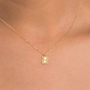 May include: A delicate gold necklace featuring a small, square pendant with a letter "B" design. The pendant is adorned with small, sparkling accents. The necklace is displayed on a person's neck, showcasing its elegant and minimalist style.