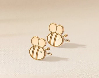 Könnte beinhalten: Ein Paar goldene Bienen-Ohrstecker mit weißen Emaille-Akzenten. Die Ohrringe befinden sich auf einem hellbeigen Hintergrund.