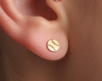 May include: A close-up of a person's ear with two gold earrings. The top earring is a small flower with black stones and a single white stone in the center. The bottom earring is a small circle with a baseball stitch design.