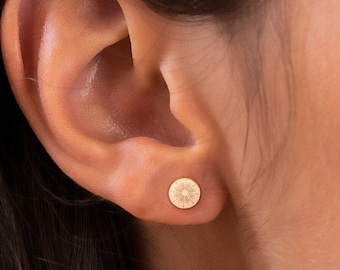 Peut inclure: Gros plan d'une boucle d'oreille dorée avec un motif de soleil. La boucle d'oreille ronde est fixée au lobe de l'oreille. L'arrière-plan montre une oreille et des cheveux.