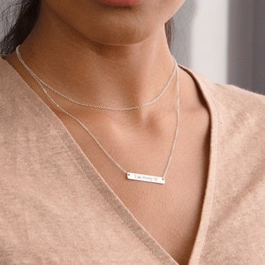 May include: A woman with dark hair wearing a light brown v-neck top and a silver necklace with a small bar pendant.