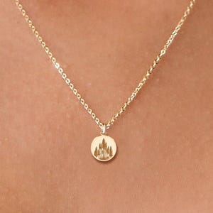 Puede incluir: Delicado collar dorado con un pequeño colgante redondo con la silueta detallada de un castillo. El collar tiene una fina cadena y se muestra sobre un fondo de tono de piel. También es visible una segunda cadena más corta.