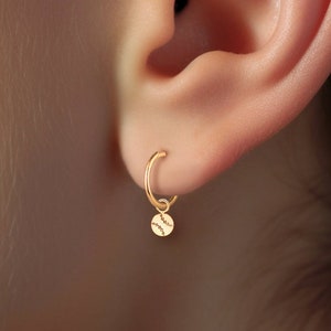 May include: Gold hoop earrings with a small, round charm hanging from the bottom. The charm has a delicate, textured design.