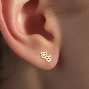 May include: A pair of gold-toned earrings featuring a smiling and a frowning face, resembling theater masks.