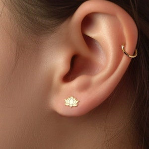 May include: A close-up of a person's ear with a small gold lotus flower stud earring. The ear also has a gold hoop earring in the upper cartilage.