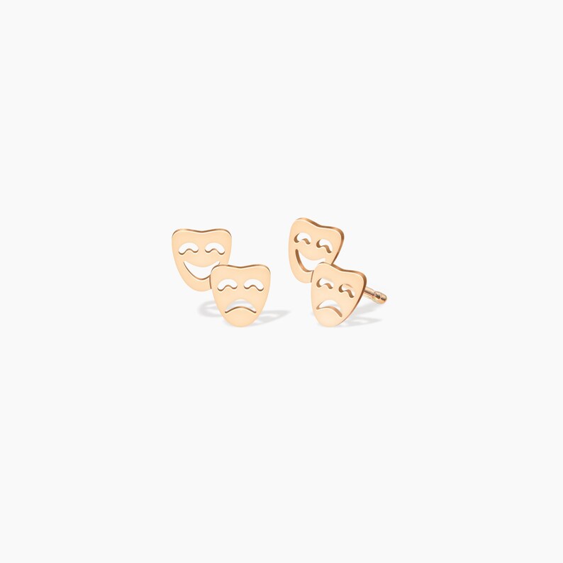 May include: A pair of gold-toned earrings featuring a happy and sad face design. The earrings are made of metal and have a post back.